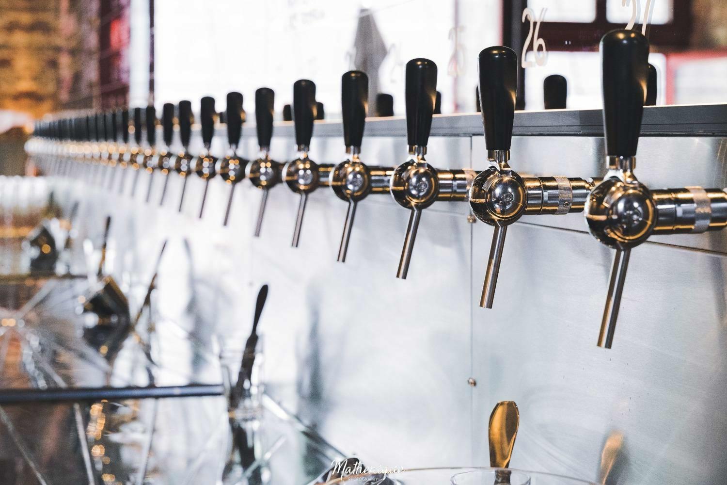 Rij van biertaps aan een glanzende toog in Bar Dragòn in Gent, met focus op de metalen tapkranen en stijlvolle baromgeving