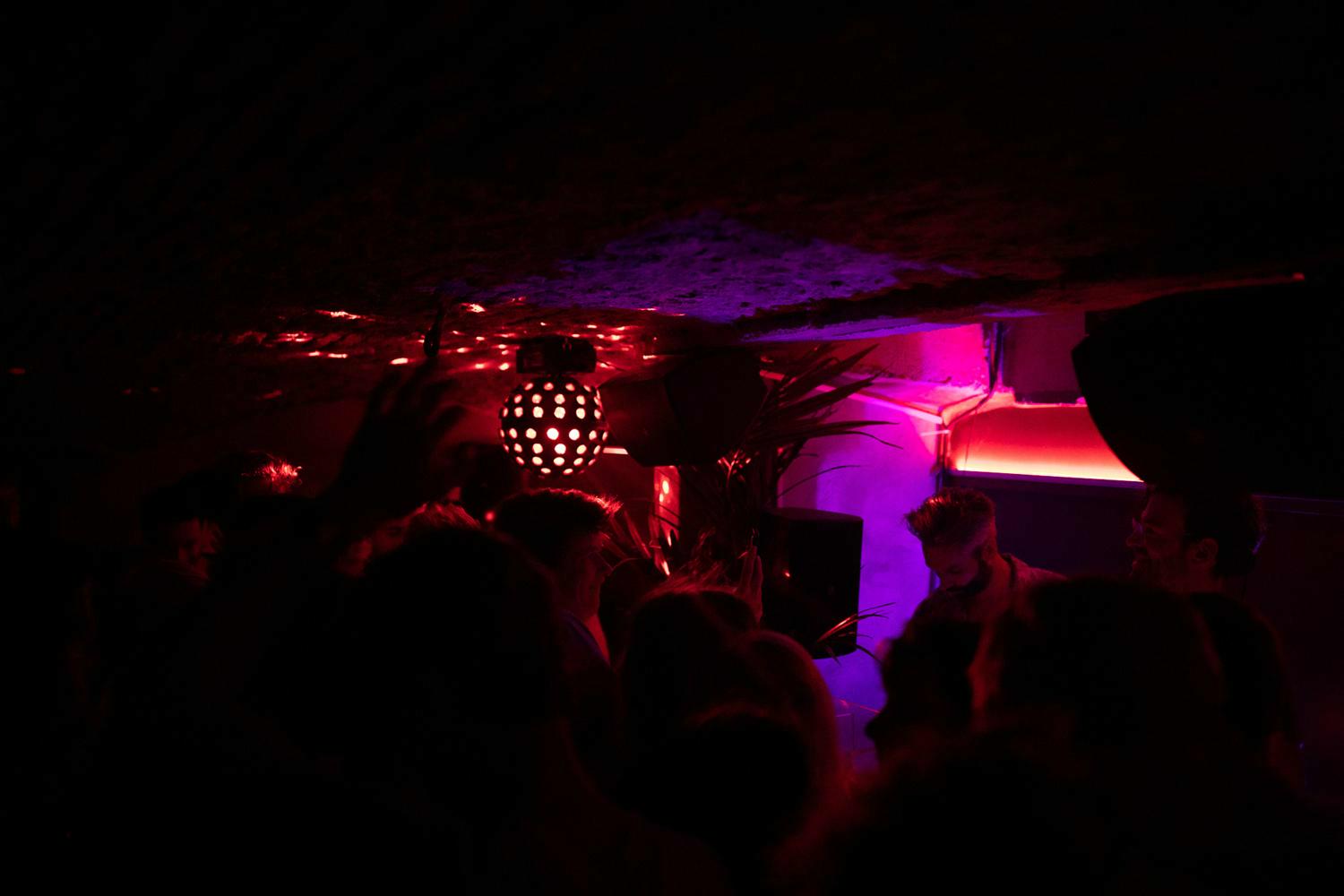 Donkere clubscène bij Bar Dragon in Gent met rood en paars licht, dansende mensen en een DJ die muziek draait onder een lage plafondverlichting.