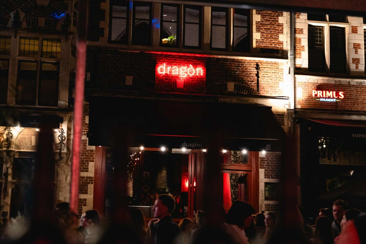 Avondscène van de façade van Bar Dragon in Gent, met een rood neonbord waarop 'dragon' staat te midden van historische bakstenen gevels. Voor de ingang staan mensen te praten en genieten van de sfeer onder warme verlichting.