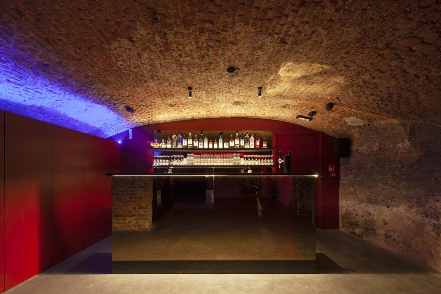 Stijlvolle kelderbar van Club Dragòn met gewelfde bakstenen plafonds, moderne verlichting in rood en blauw, en een glanzende toog met flessen drank en glazen op de achtergrond.